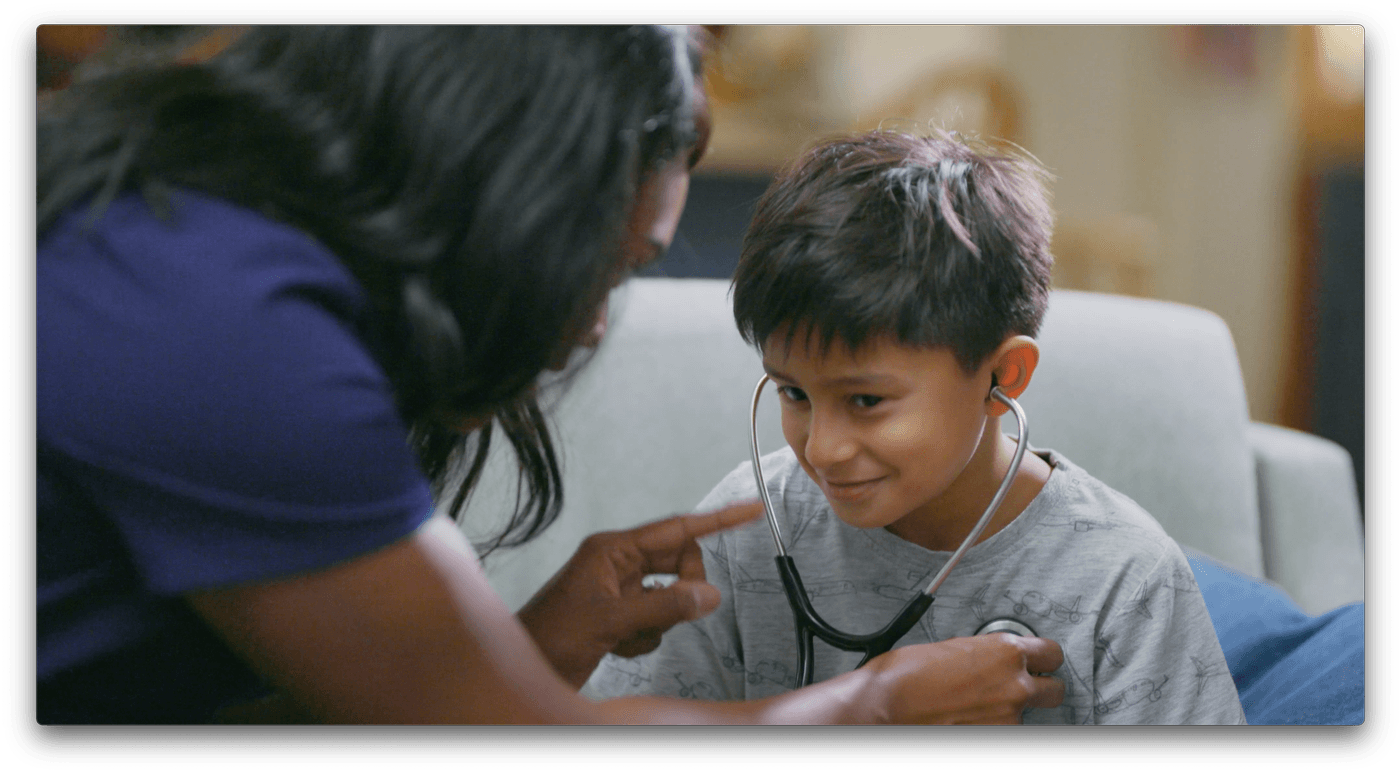 A healthcare worker wearing a dark blue uniform sits on a sofa and uses a stethoscope to examine a young boy who is smiling while wearing the earpieces.