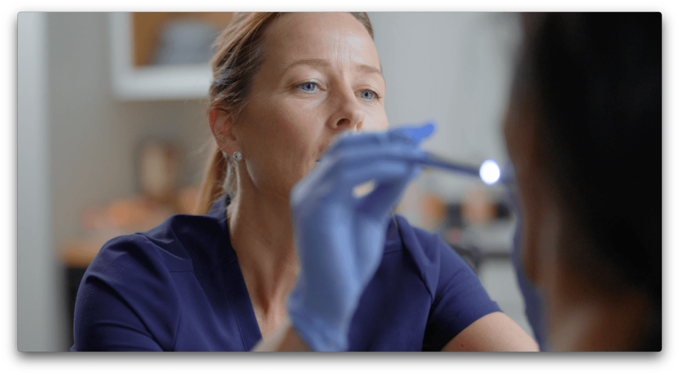 A female medical professional wearing blue gloves uses a small penlight to examine a patient's mouth in a clinical setting.