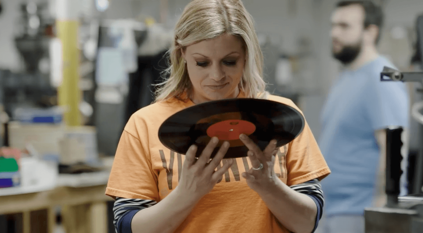 A woman with blonde hair wearing an orange t-shirt examines a vinyl record in her hands, while a blurred man stands in the background of the workshop.