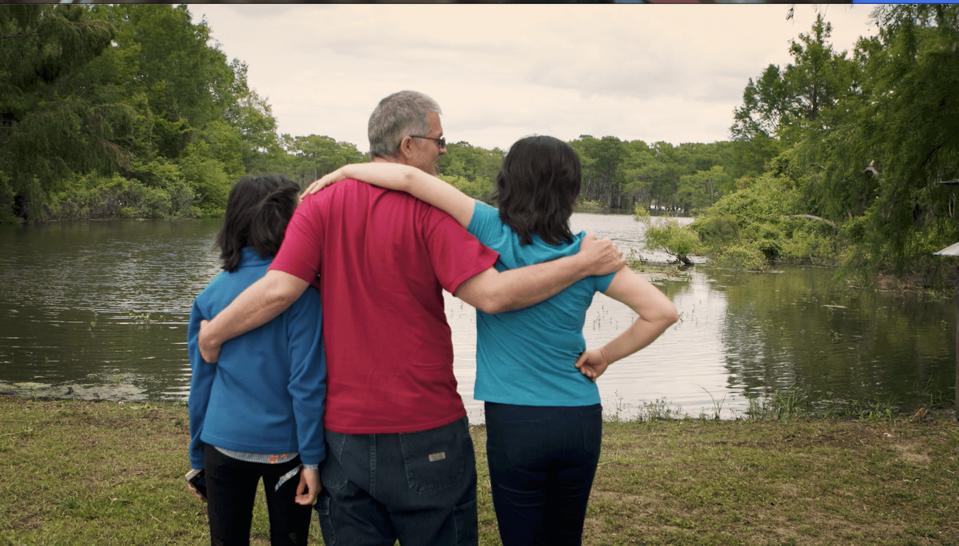 A man and two people stand with their backs to the camera, arms around each other, looking out at a calm lake surrounded by lush green trees.