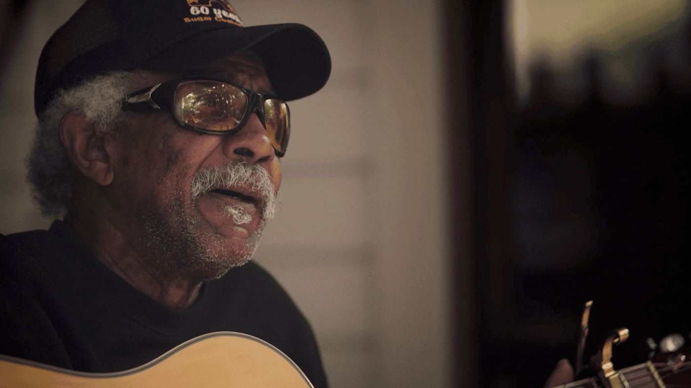 An elderly Black man with a graying mustache and afro, wearing a baseball cap and tinted sunglasses, sings while playing an acoustic guitar.