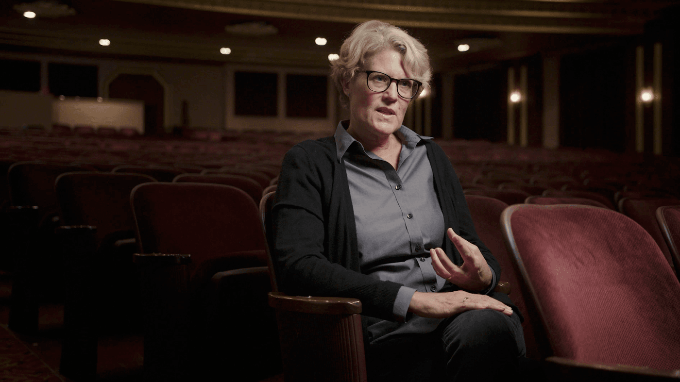 A middle-aged woman with short blonde hair and glasses sits in a theater chair, gesturing while speaking to someone off-camera.