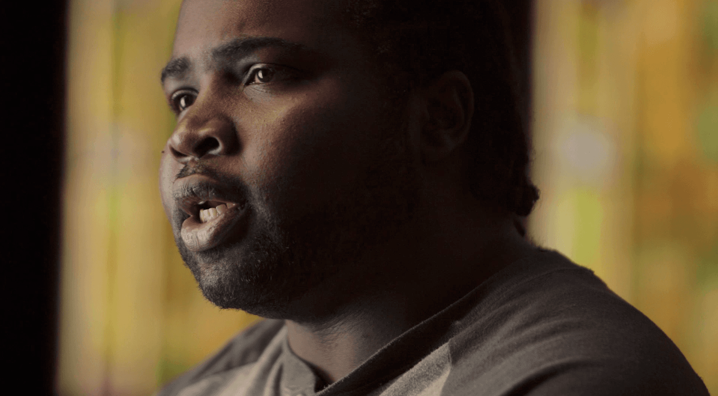 A close-up profile shot captures a man with dark skin and a beard, looking slightly upward while speaking against a soft, out-of-focus background with warm yellow light.
