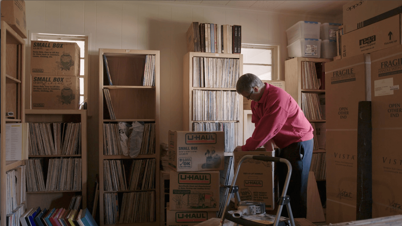 A man in a red sweater packs vinyl records into a U-Haul cardboard box within a room filled with floor-to-ceiling wooden shelving units densely packed with record albums.