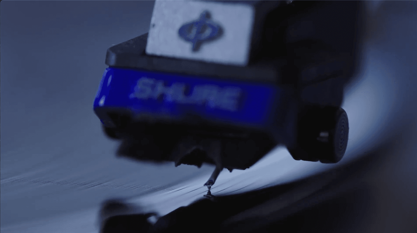 A close-up, low-angle shot of a Shure phono cartridge with a blue faceplate tracking a vinyl record, with its needle resting precisely in the groove.