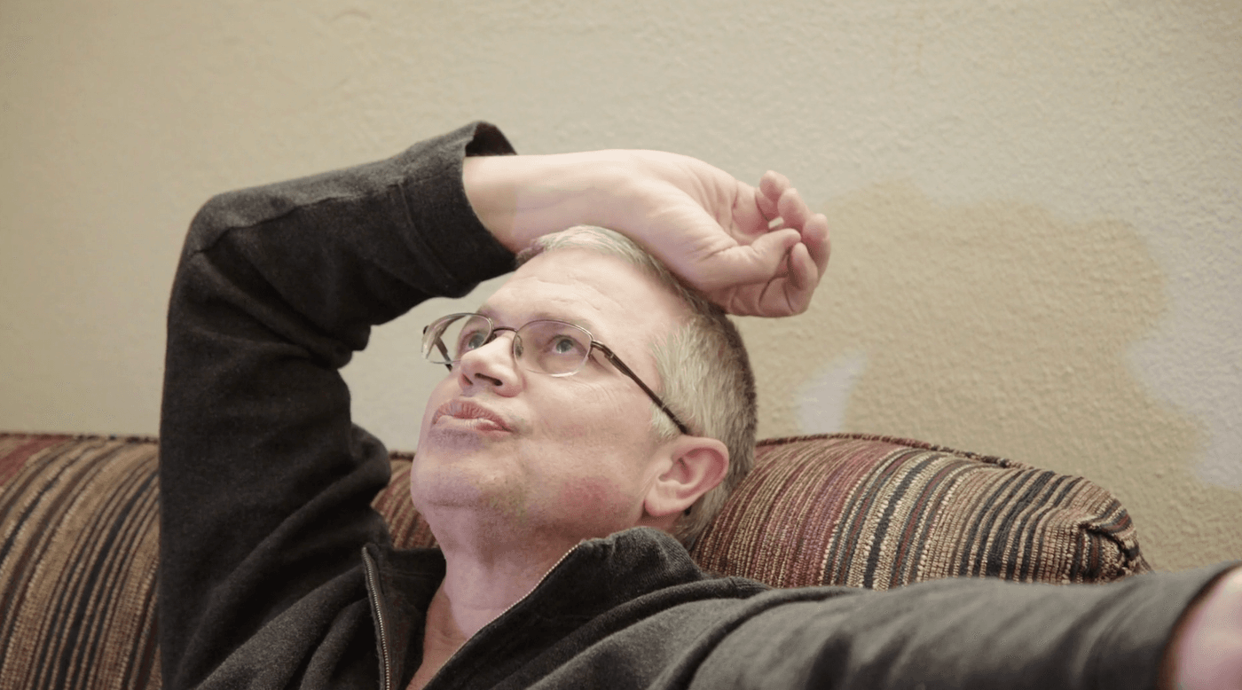 A man wearing glasses and a dark zip-up jacket sits on a striped couch with his hand resting on his forehead while looking upward.