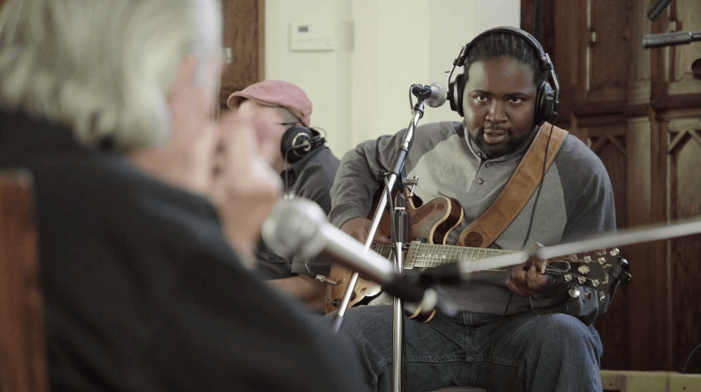 A focused man with locs wears headphones while playing a semi-hollow body guitar in a studio, sitting near a musician with a red cap and a foreground figure with silver hair.