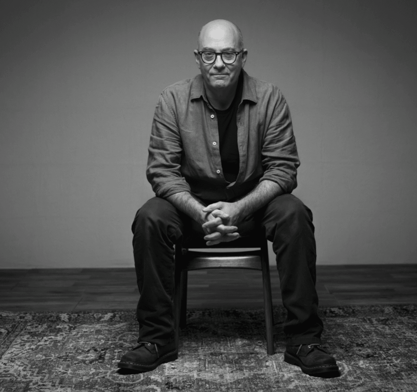 A black-and-white portrait of a bald, bespectacled man wearing a button-down shirt over a t-shirt, sitting with his hands clasped on his lap on a wooden chair atop a patterned rug.