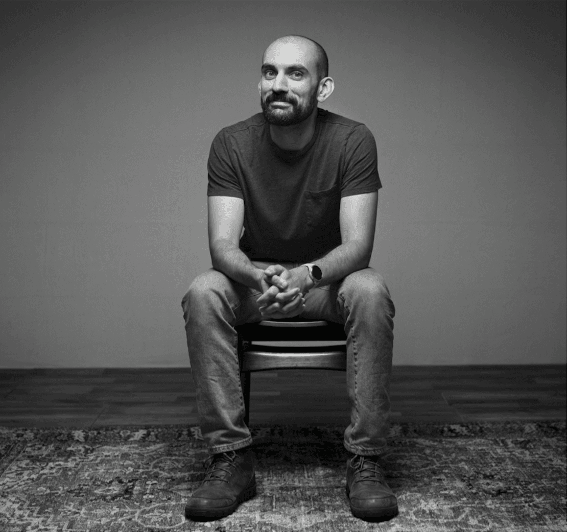 A black-and-white portrait of a man with a beard and shaved head, wearing a t-shirt and jeans, sitting on a chair with his hands clasped in his lap against a plain background.