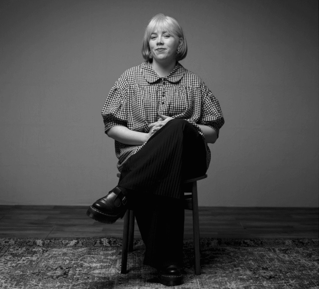 A blonde woman with a bob hairstyle sits on a stool wearing a gingham-patterned collared shirt and black trousers, posing with her legs crossed against a plain studio background.