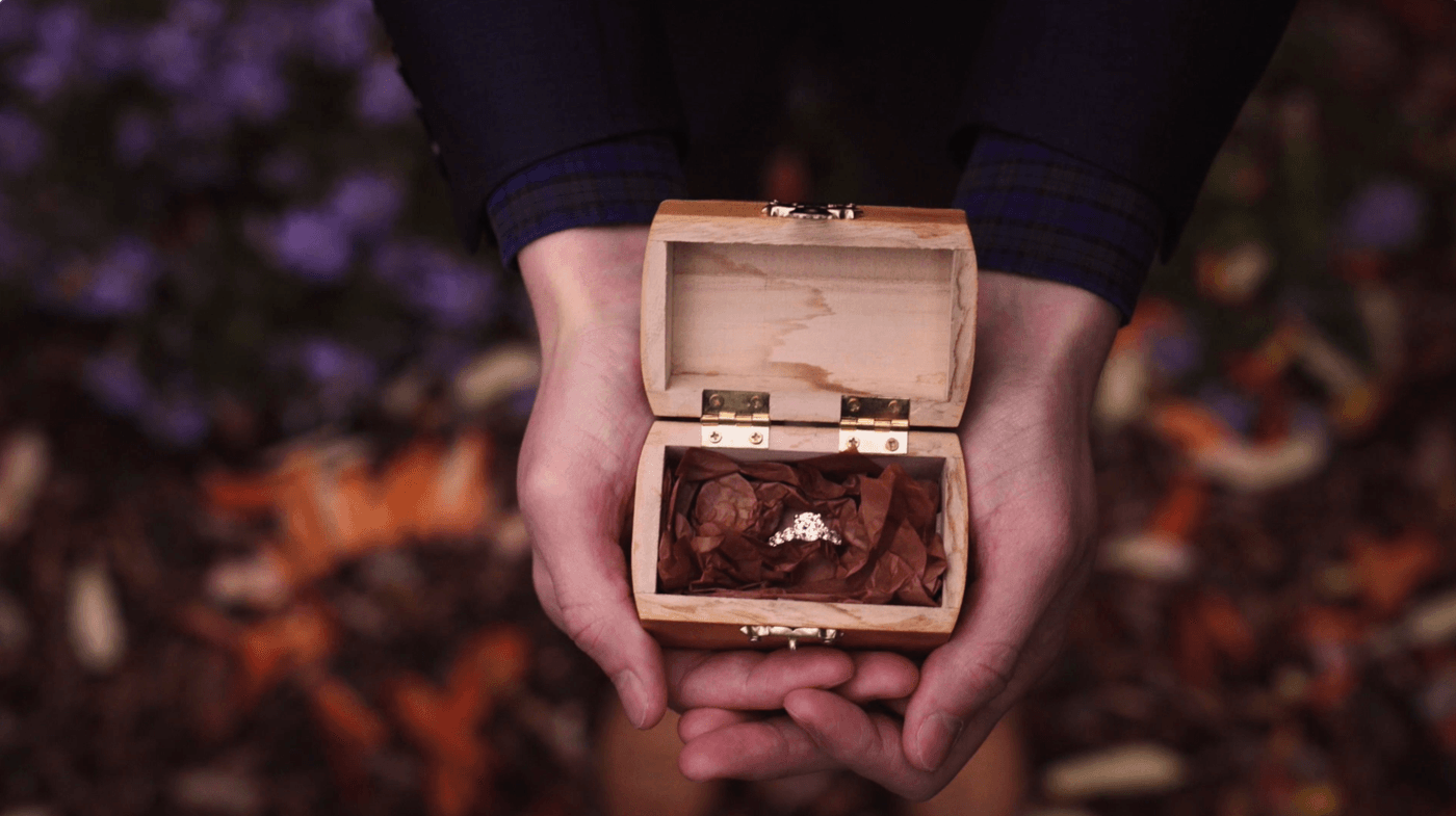 A person in a dark blue blazer with a plaid shirt cuff holds an open, small wooden box containing a diamond engagement ring nestled in brown tissue paper against a background of blurred autumn leaves.