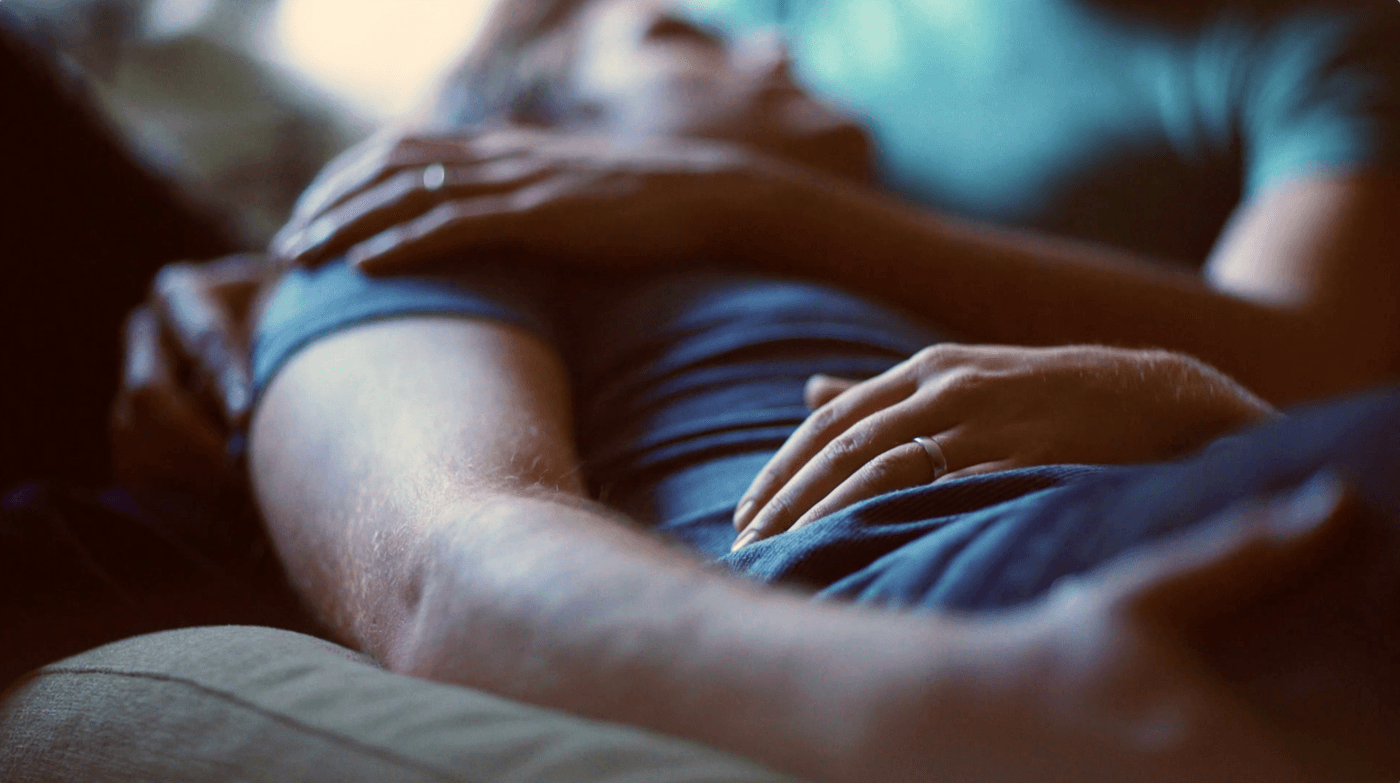 Close-up shot of two people cuddling while lying down, focusing on their hands resting on each other and wearing wedding bands.