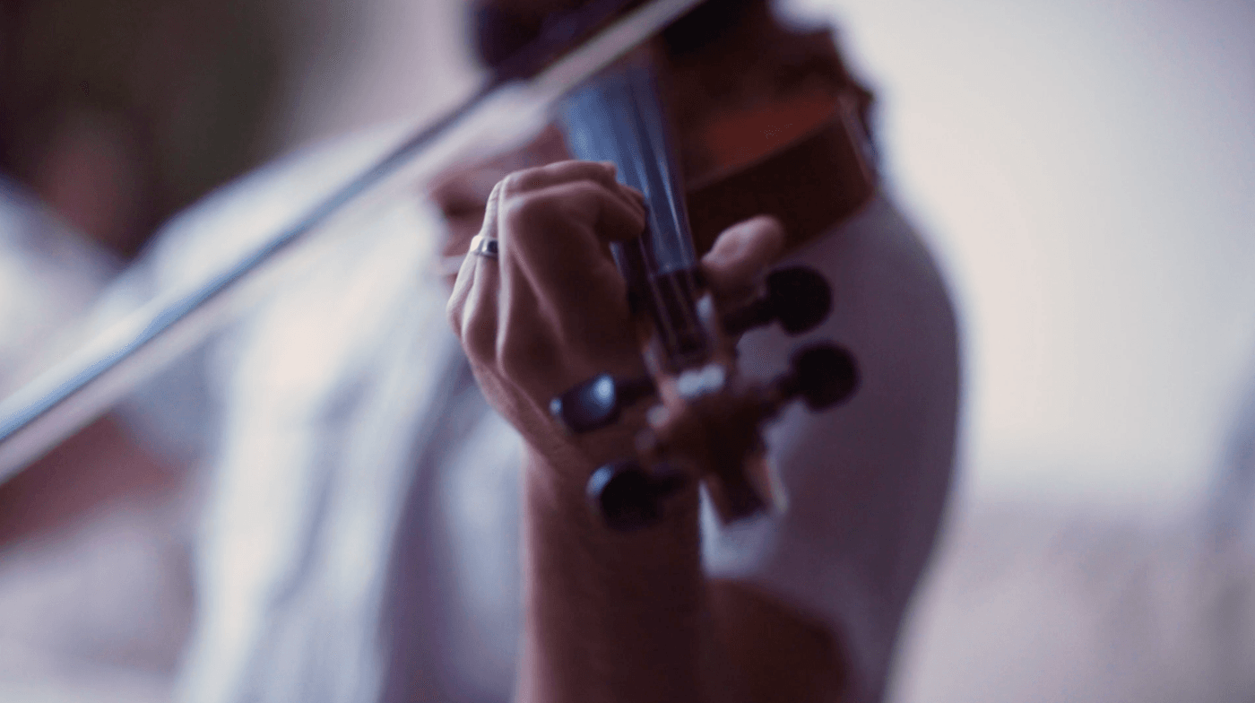A close-up, shallow-depth-of-field view of a person's hand wearing a silver ring while fingering the neck of a violin, with a blurred bow extended across the frame.