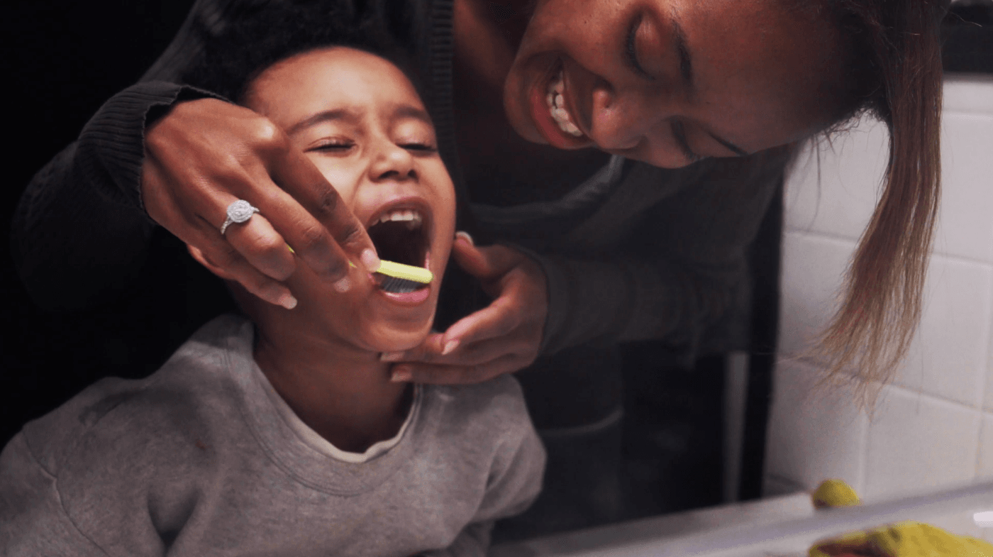 A mother with a smiling expression helps her young son brush his teeth in a bathroom.