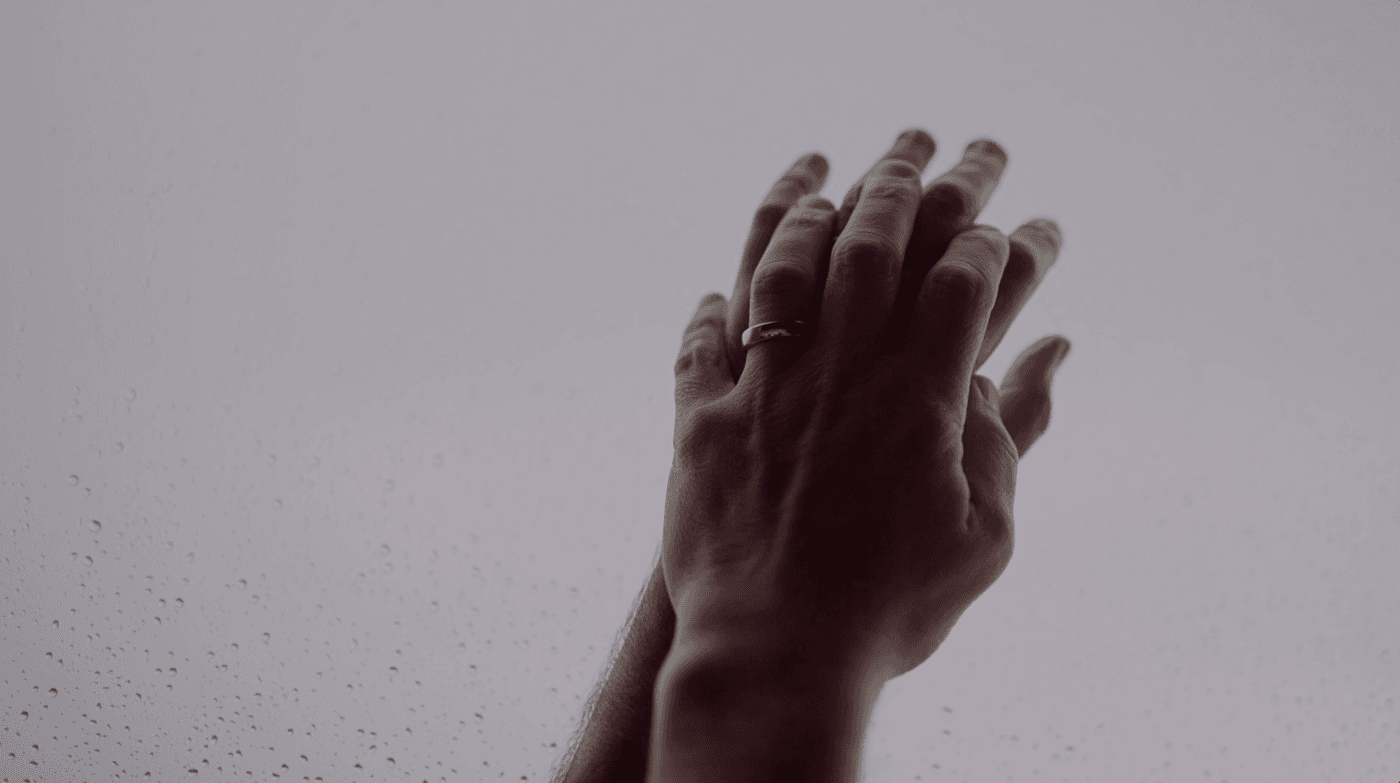 A close-up view of two hands reaching upward toward a light-filled, rain-streaked window, with a silver band visible on one of the fingers.