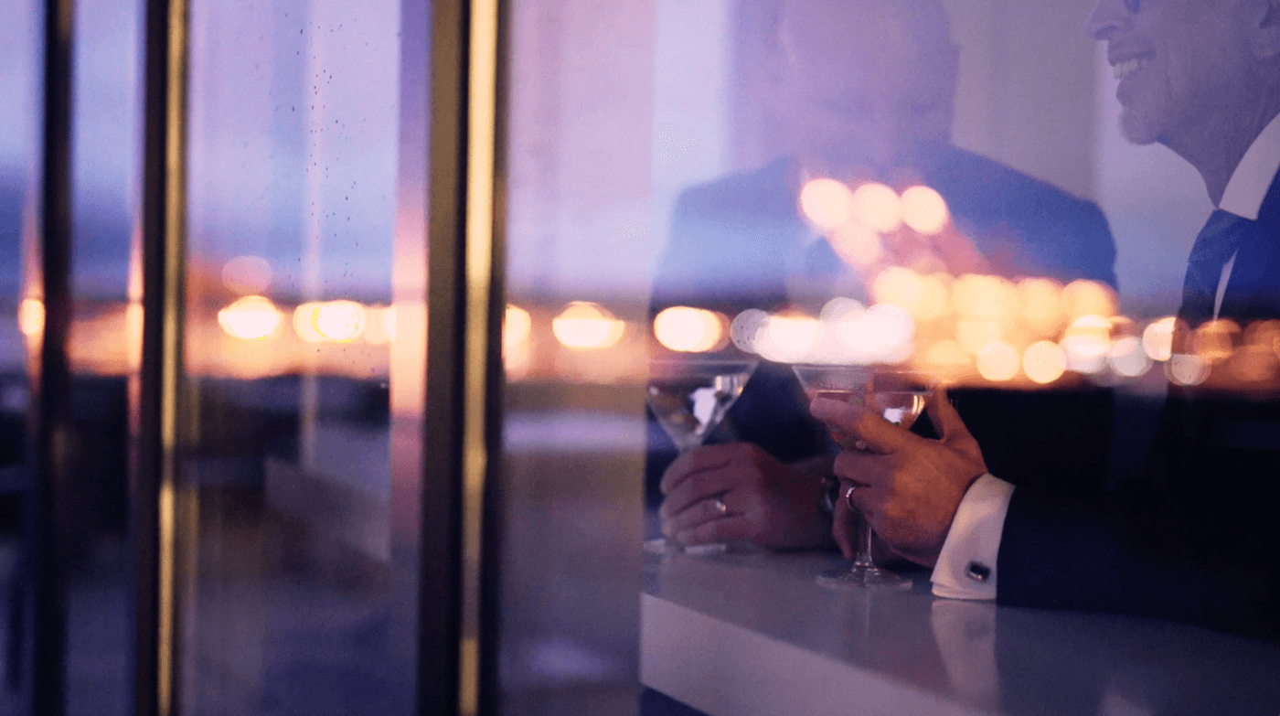 A side view, reflected through a window, shows two men in suits holding martini glasses at a bar with blurry city lights in the background.