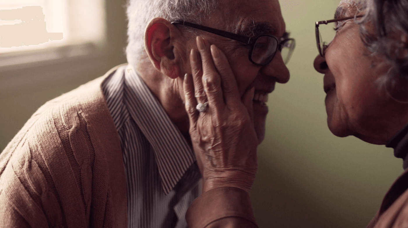 An elderly woman wearing a diamond ring gently caresses the cheek of an older man who is smiling warmly while both wear glasses.