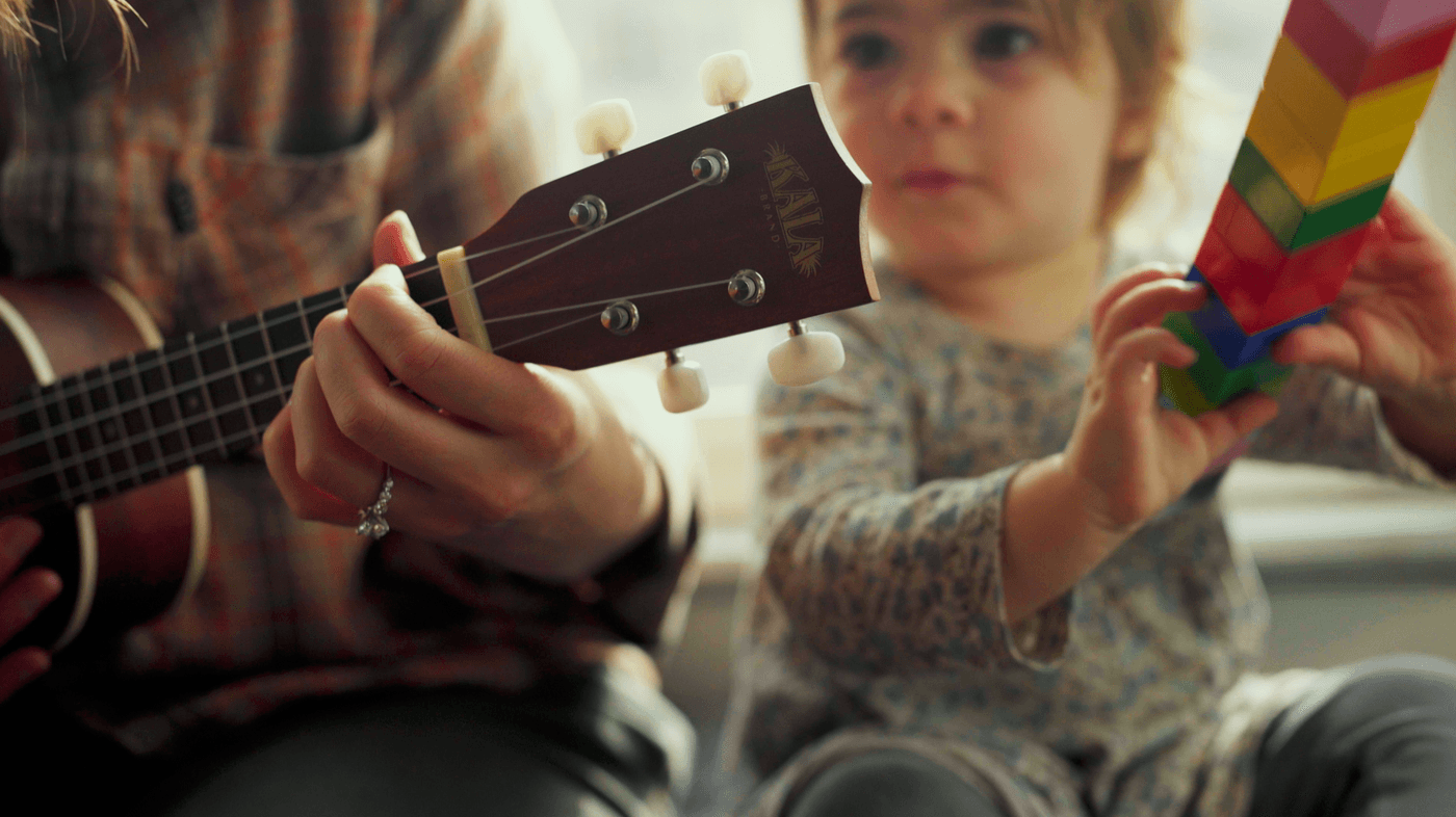 A woman with a ring on her finger plays a Kala brand ukulele while a young child sits beside her holding a stack of colorful building blocks.