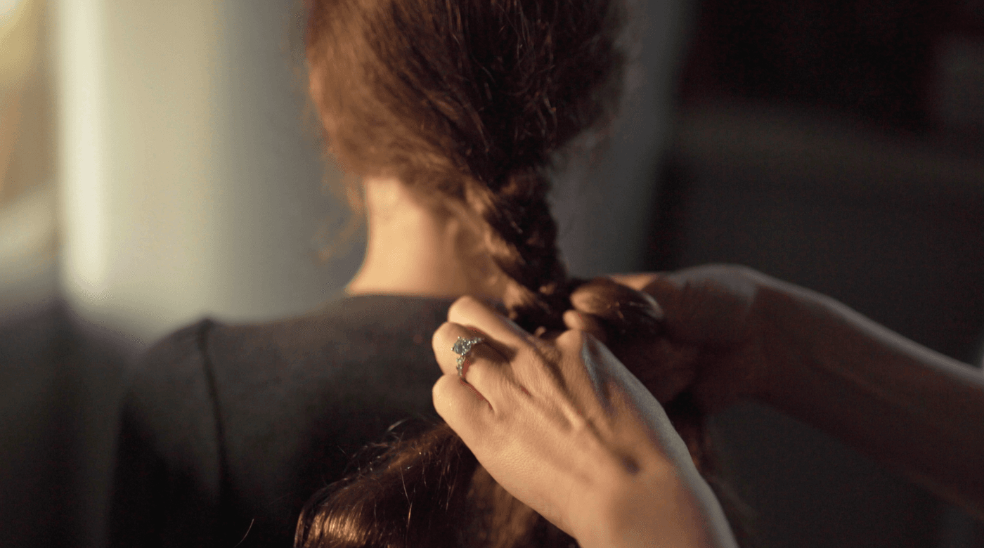 A person with a diamond ring on their finger braids the long brown hair of another person in a dimly lit, soft-focus setting.