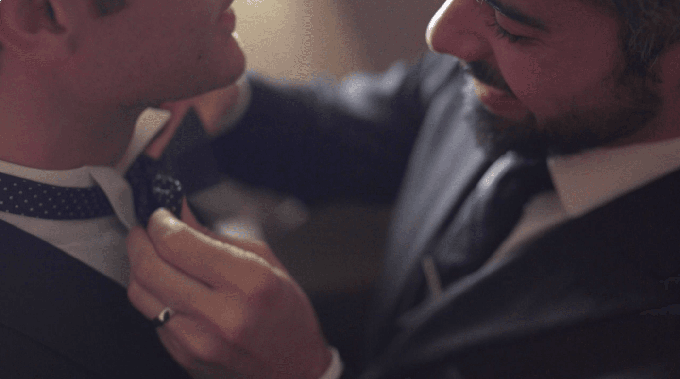A close-up shot captures a man in a black suit with a beard, smiling warmly as he adjusts the polka-dot bow tie of the man standing in front of him.