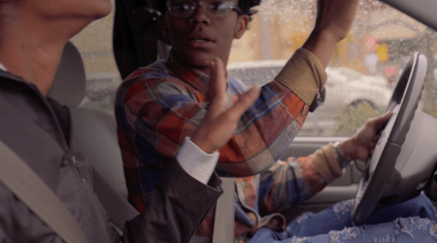 A person in a plaid shirt with glasses sits in the driver's seat of a car while speaking to another person in the passenger seat, with rain visible on the car window.