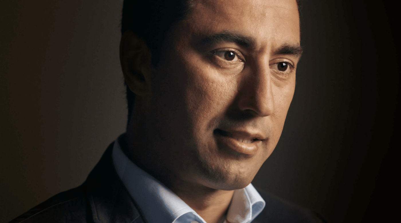 A medium close-up, profile-oriented portrait of a man with dark hair and brown eyes wearing a light blue dress shirt and a dark blazer against a dark, moody background.