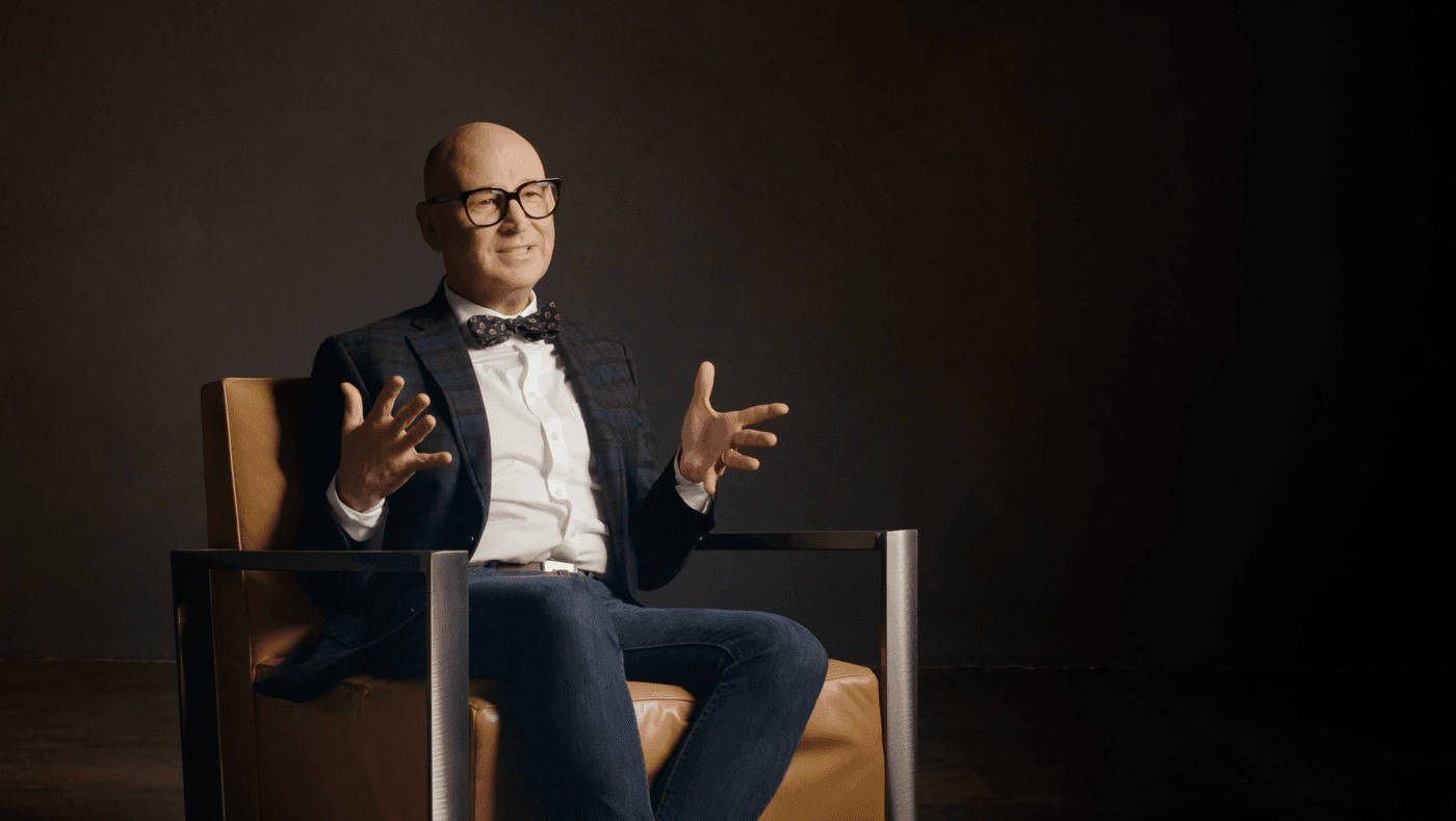 A bald man with glasses and a bow tie wears a plaid blazer and jeans while sitting in a mustard-colored armchair and gesturing with both hands against a dark background.