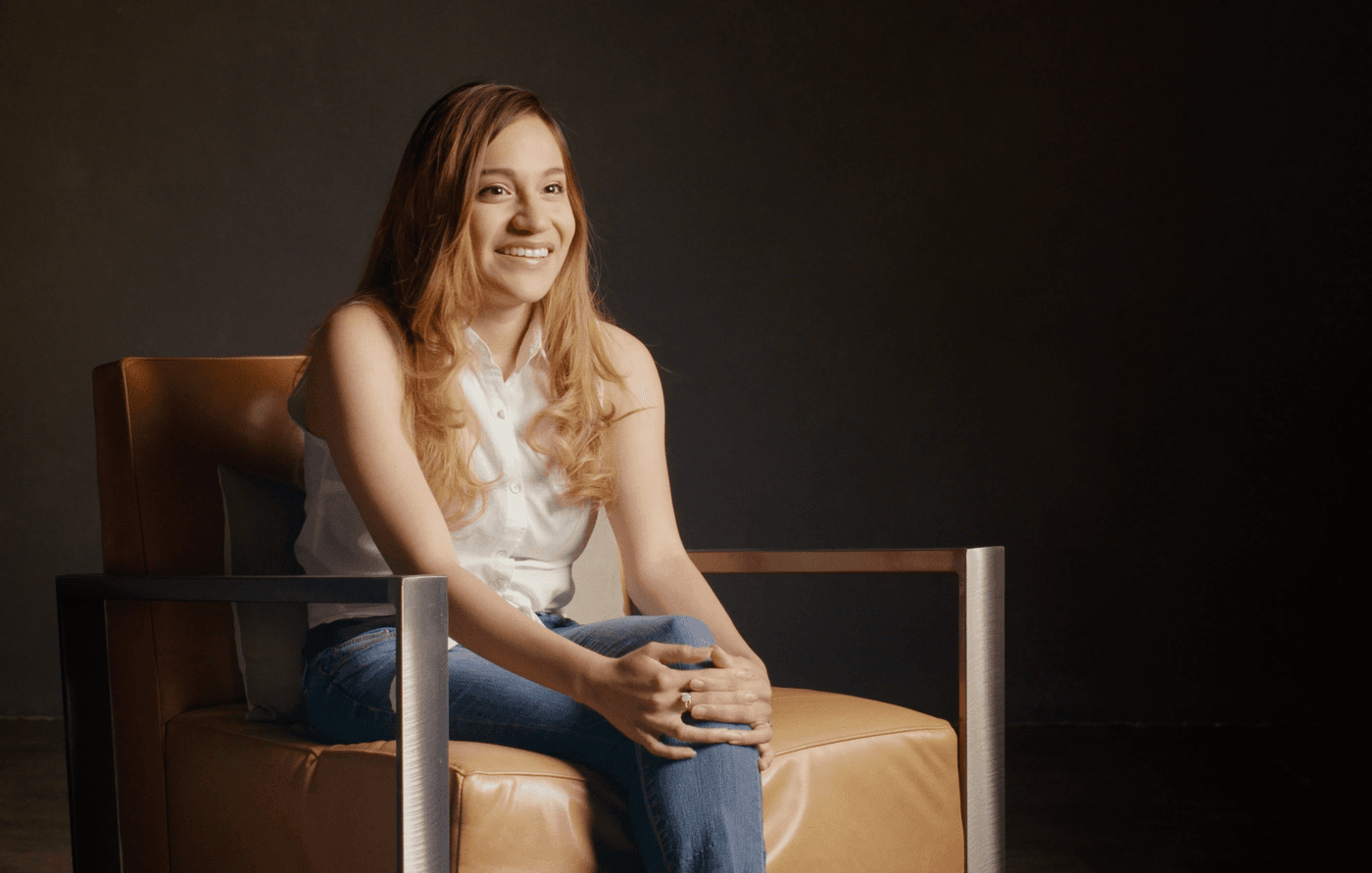 A woman with long light brown hair smiles while sitting in a modern leather chair against a dark, plain background.
