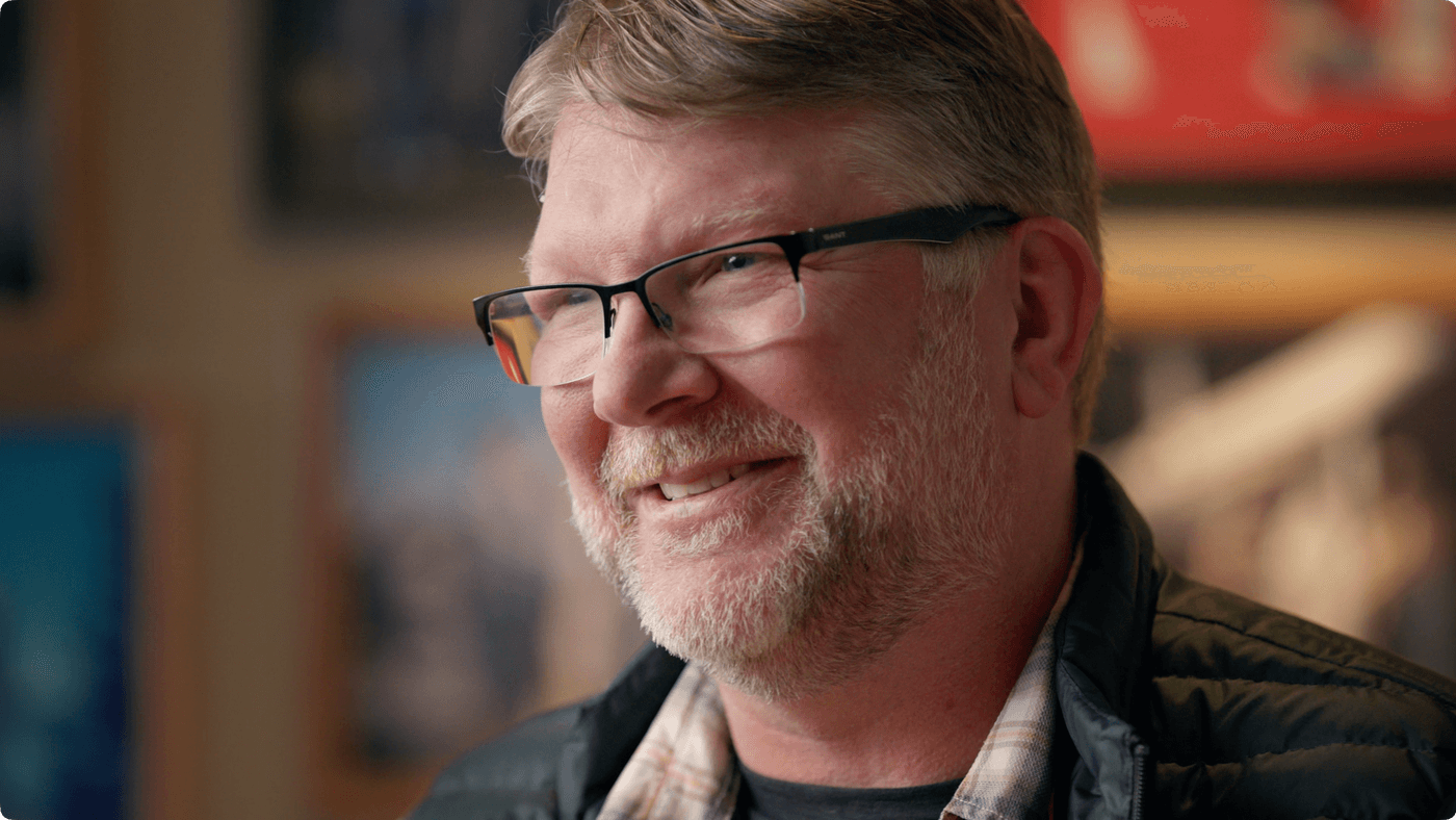 A middle-aged man with short, light-brown hair and a light beard wears black-rimmed glasses, a plaid collared shirt, and a black quilted jacket while smiling slightly in a softly lit room.