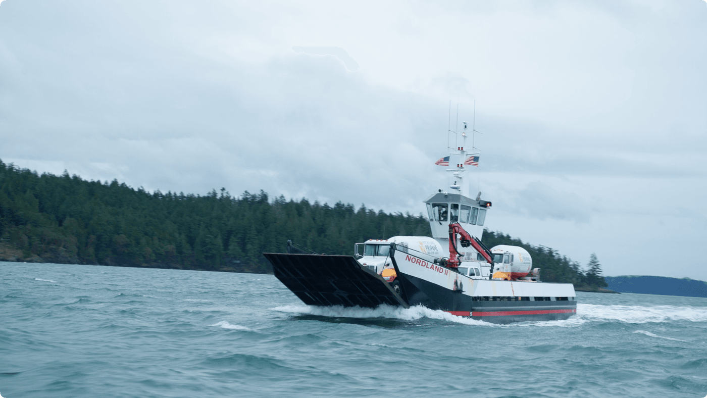A white and black landing craft ferry named Nordland II transports a propane tanker truck across choppy water, with a forested coastline in the background under a cloudy sky.