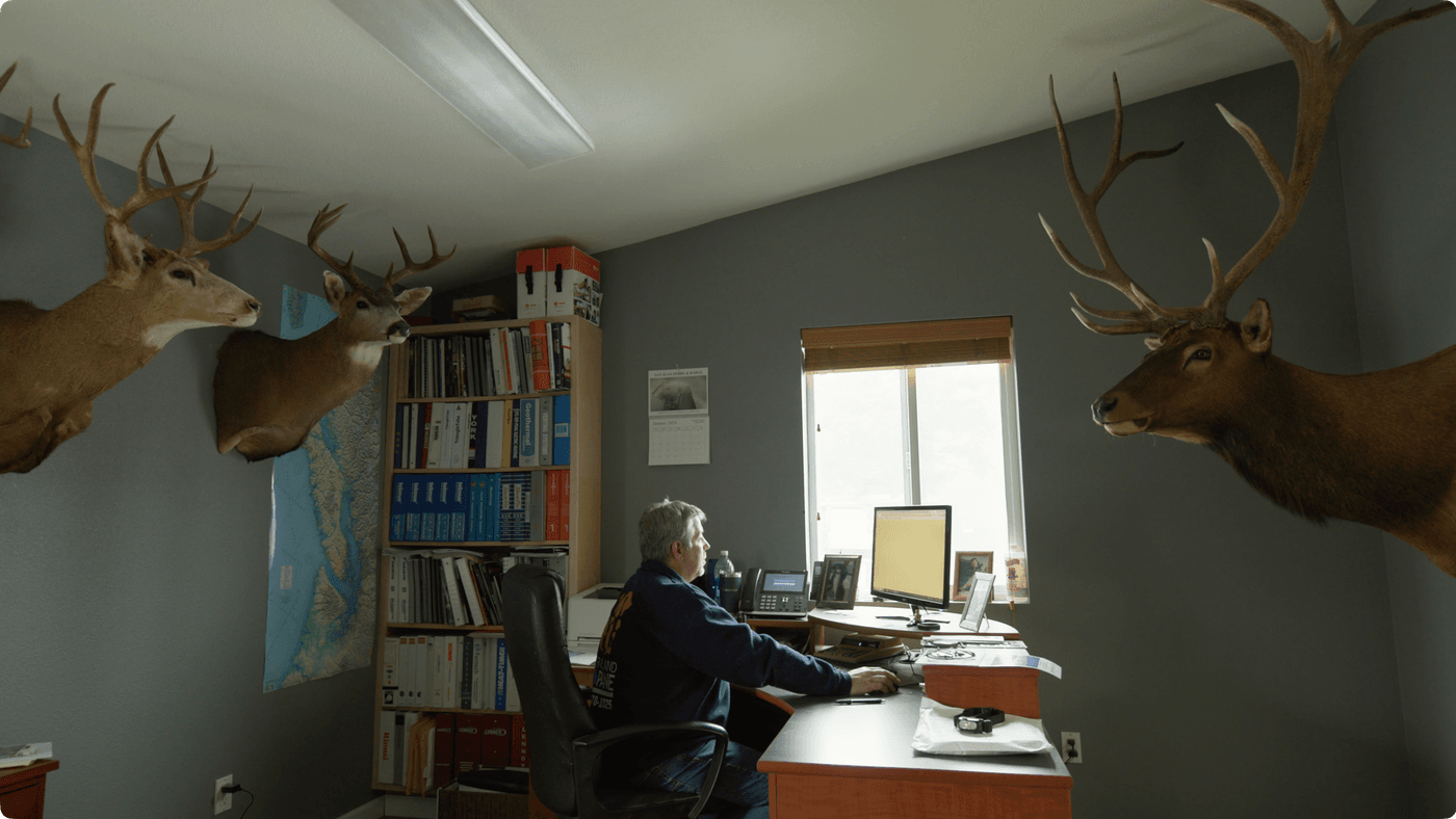 A man sits at a desk in an office adorned with mounted deer and elk heads, working on a computer near a window and a bookshelf filled with binders.