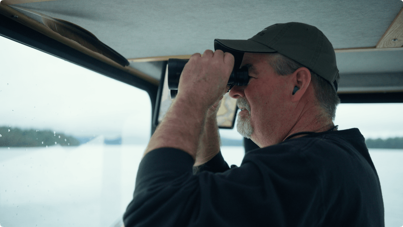 A middle-aged man with a graying beard and green baseball cap looks through binoculars while standing inside the cabin of a boat overlooking a calm, misty lake.