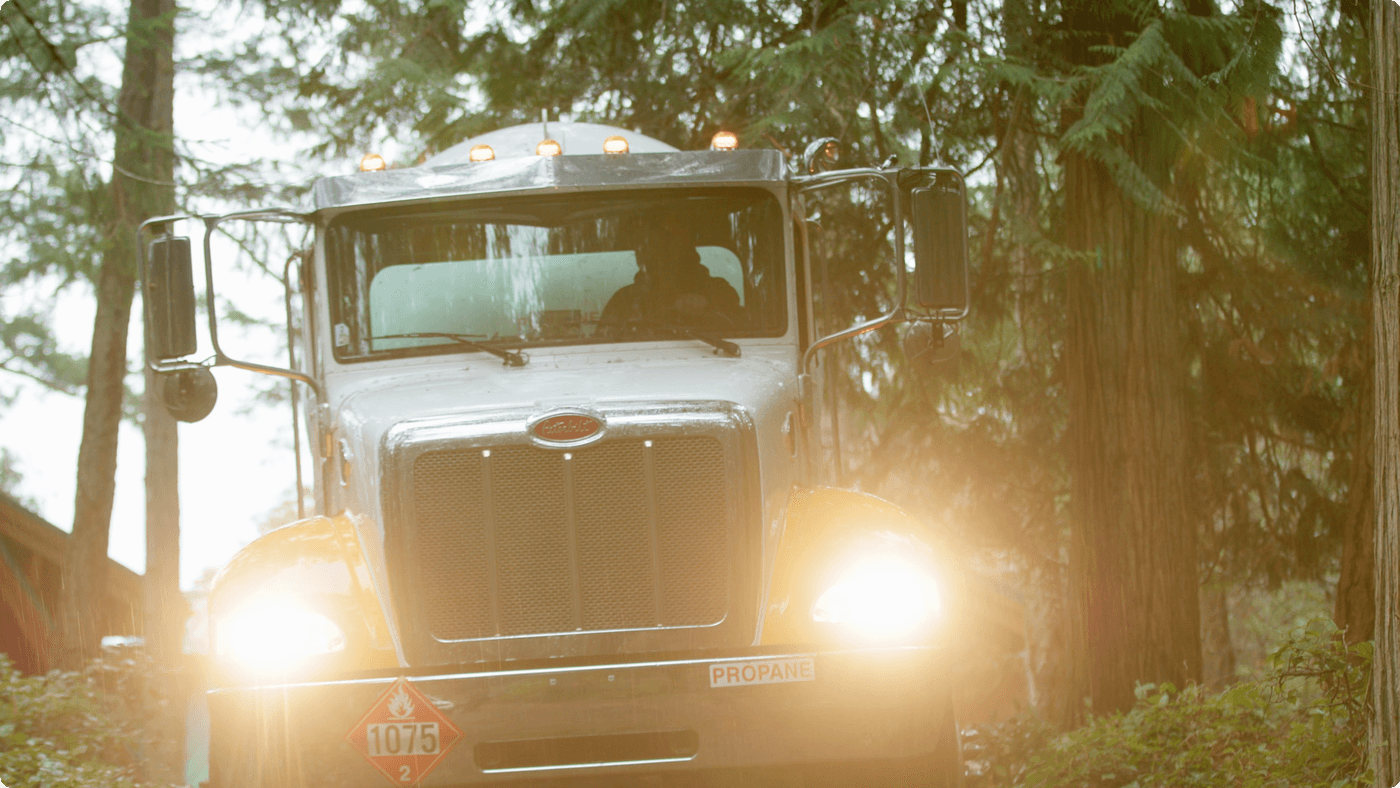 A white Peterbilt propane delivery truck with bright headlights driving through a sun-dappled, wooded area.