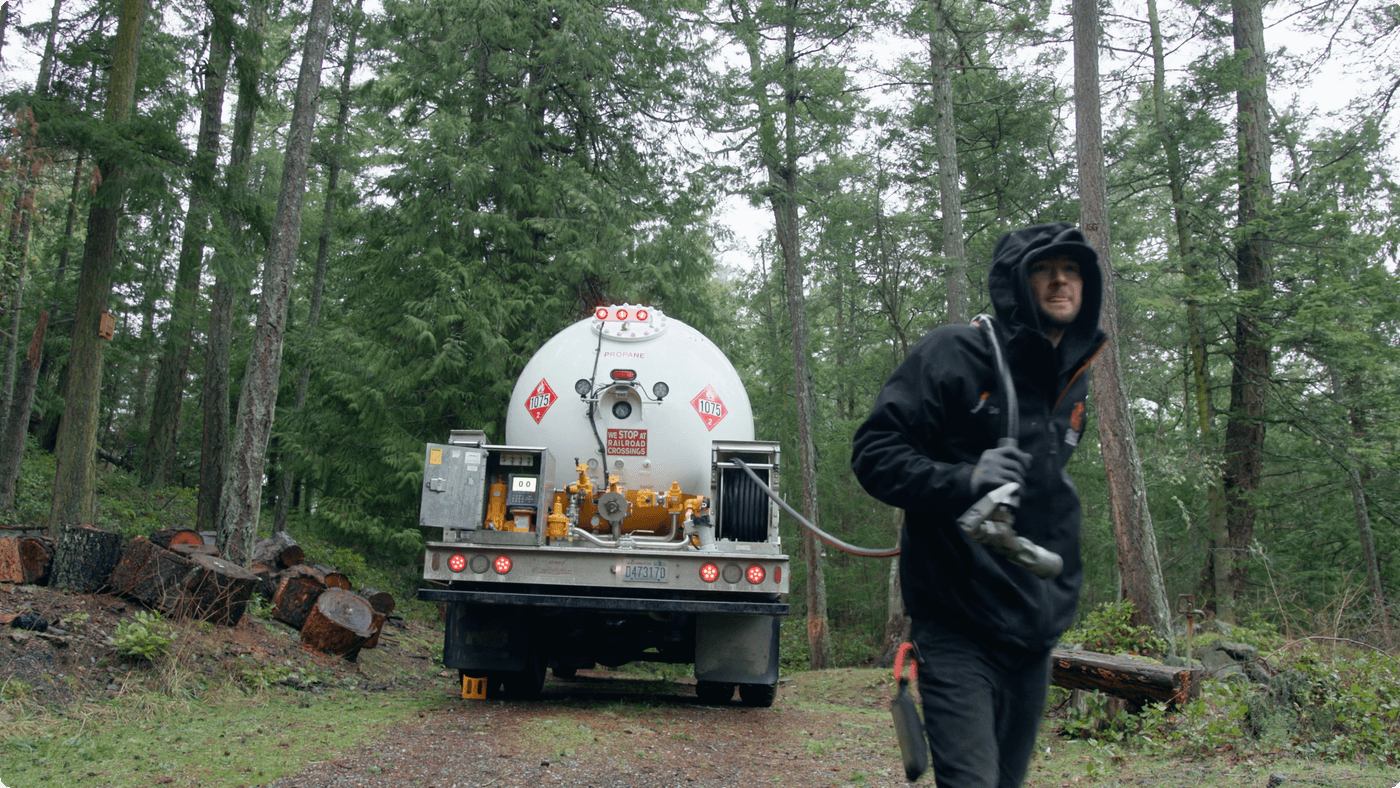A propane delivery truck is parked on a wooded gravel road while a worker in a black jacket and hood walks away from the back of the vehicle carrying a fuel hose nozzle.