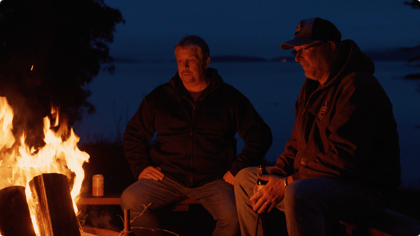 Two middle-aged men sit on a bench by a campfire at dusk, with one holding a drink and the ocean visible in the background.