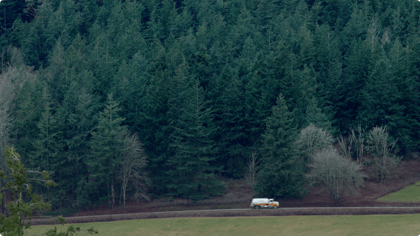A tanker truck drives along a rural road bordered by a dense evergreen forest and an open grassy field.