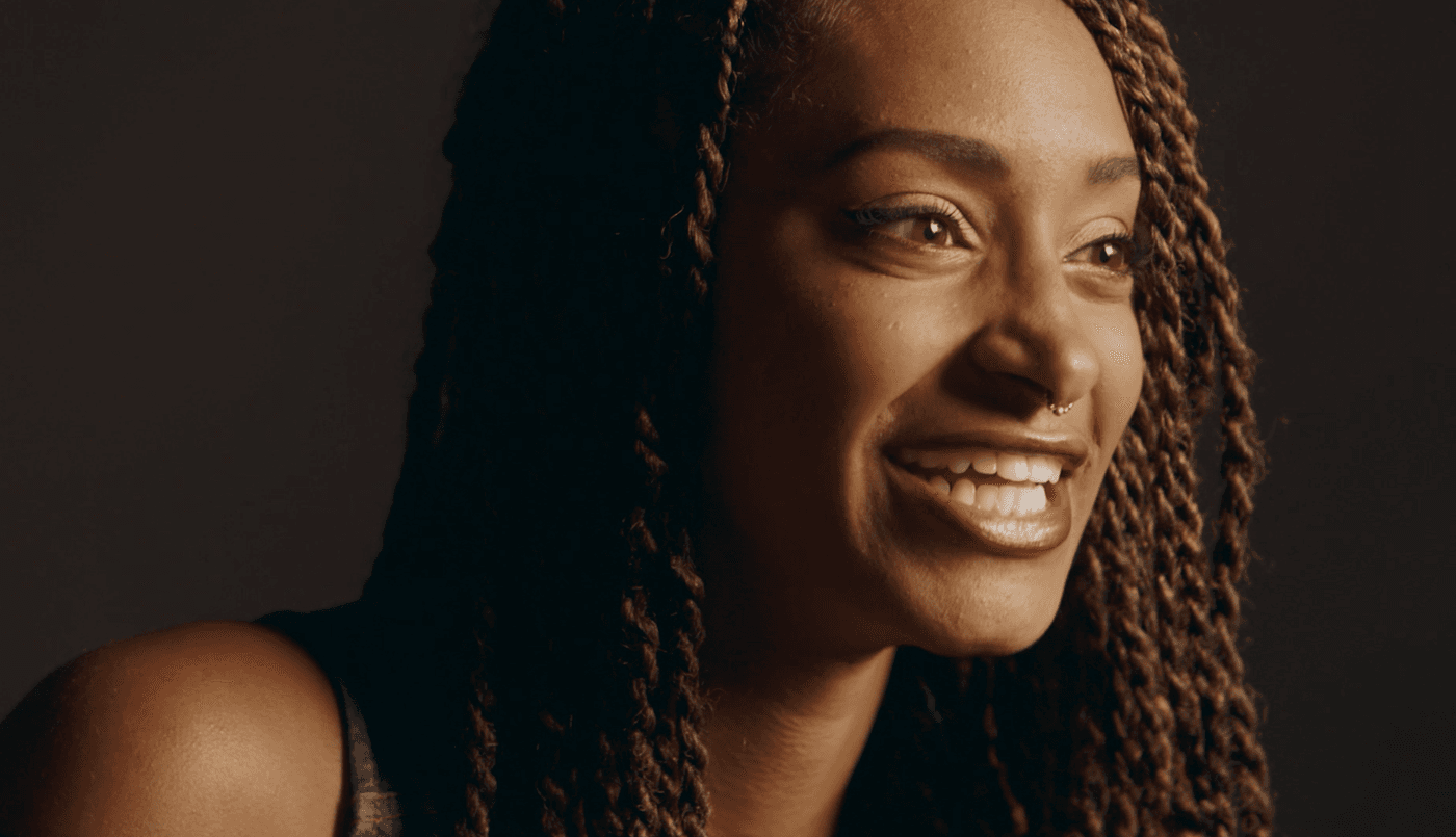 A close-up shot of a smiling woman with long, brown, braided hair, a septum piercing, and warm lighting against a dark background.