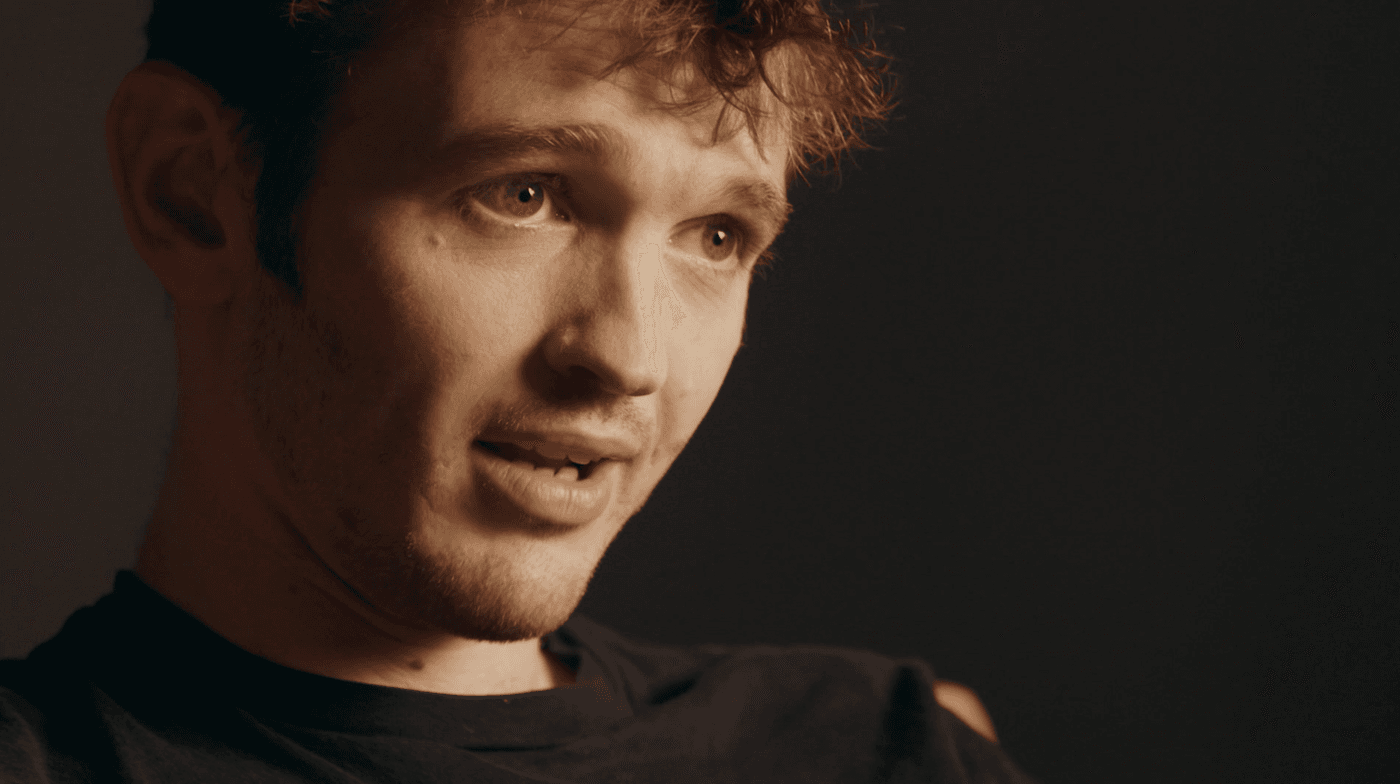 A close-up shot of a young man with curly brown hair looking off-camera to the right while speaking against a dark background.
