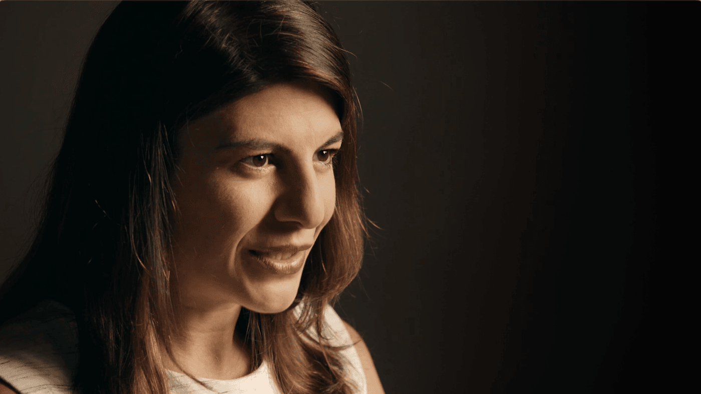 A side profile close-up shot of a woman with long brown hair, wearing a white shirt against a dark, moody background, looking slightly off-camera.