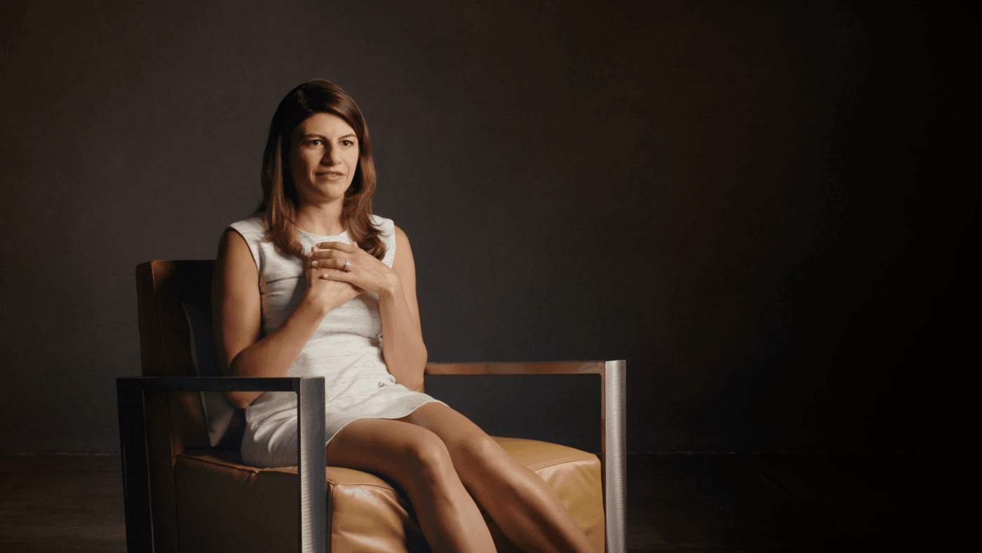 A woman with shoulder-length brown hair wears a white sleeveless dress while sitting in a tan leather armchair against a dark background.