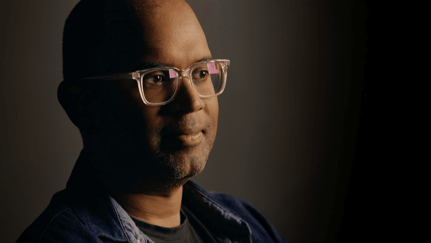 A medium close-up shot of a Black man with short hair and stubble, wearing clear-framed glasses and a dark denim jacket, looking thoughtfully toward the right against a dark, minimalist background.