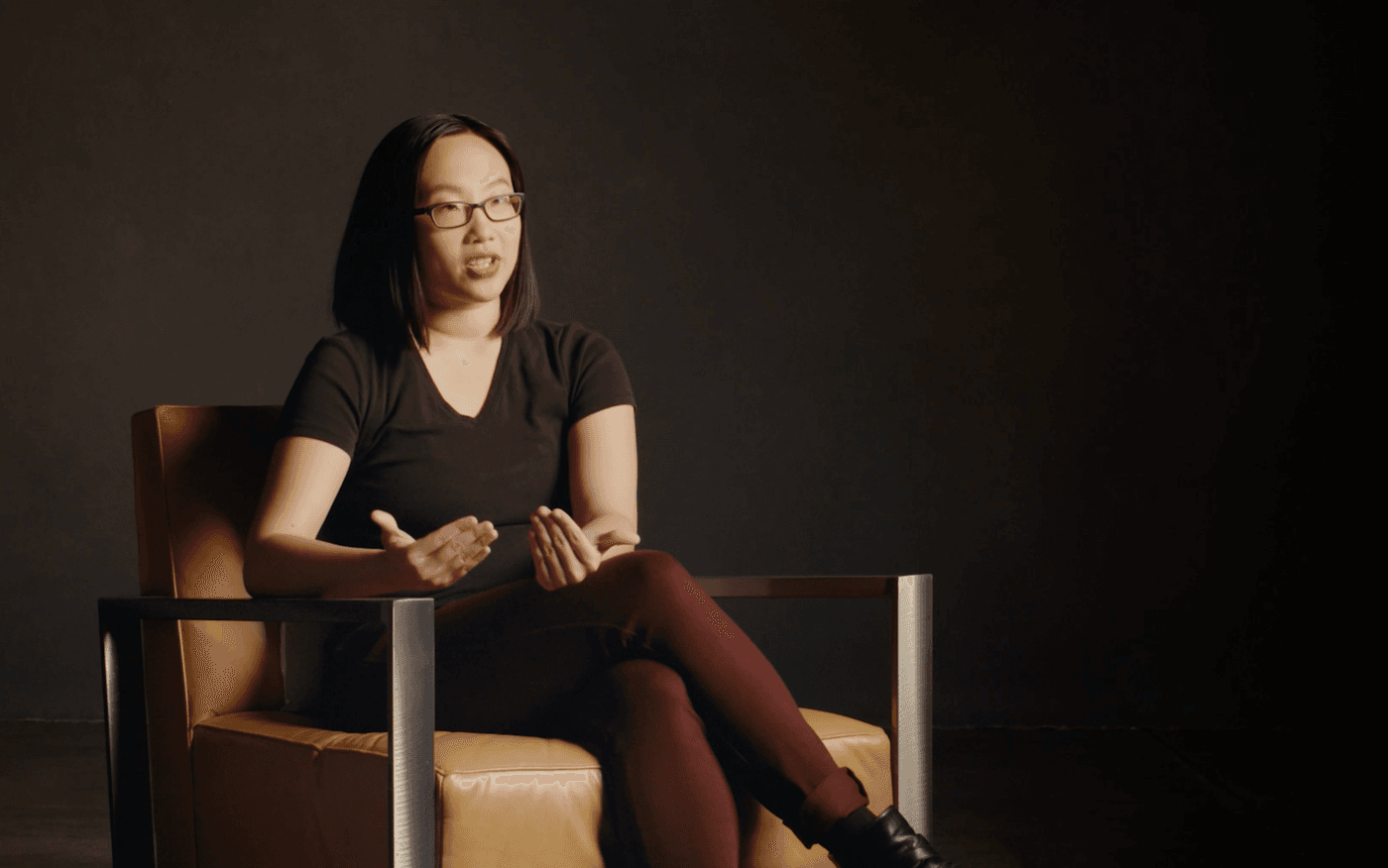 A young woman with shoulder-length dark hair and glasses sits in a tan leather chair against a dark background, wearing a black v-neck t-shirt and burgundy pants while gesturing with her hands.