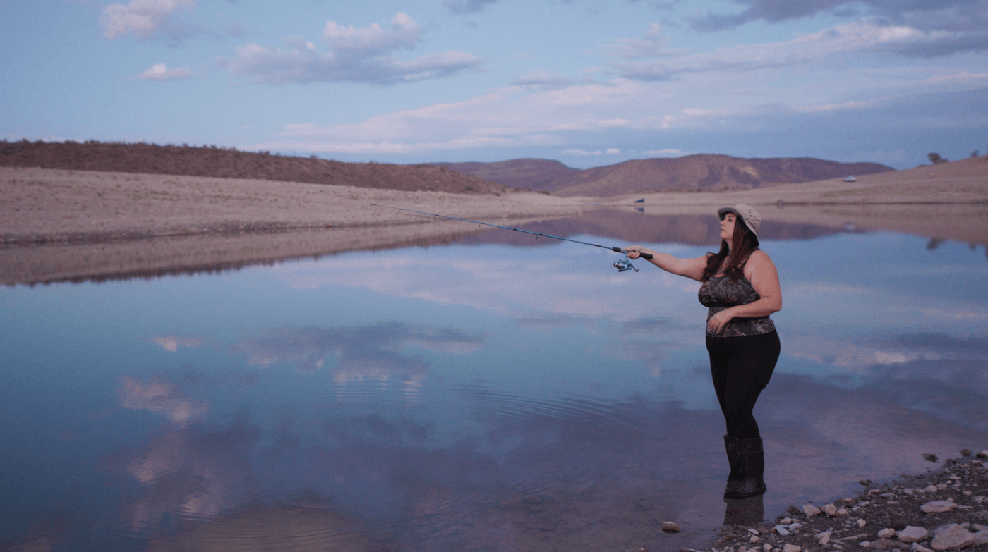 A woman wearing a camouflage tank top, leggings, and a bucket hat stands in shallow water at the edge of a lake, holding a fishing rod over the reflective surface under a twilight sky.