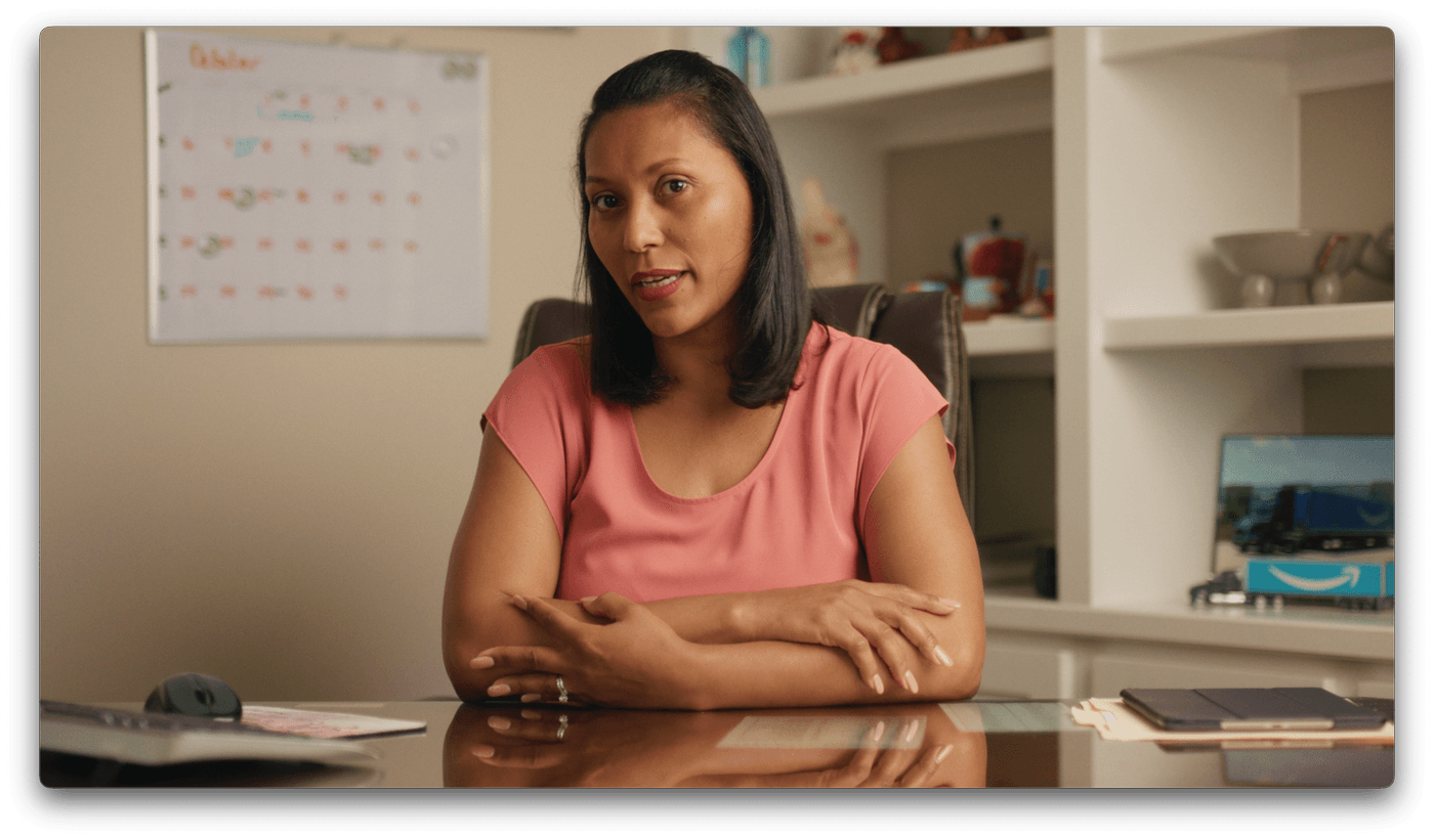 A woman with dark hair wearing a pink shirt sits at a desk with her arms crossed, positioned in front of a white shelf and a calendar on the wall.
