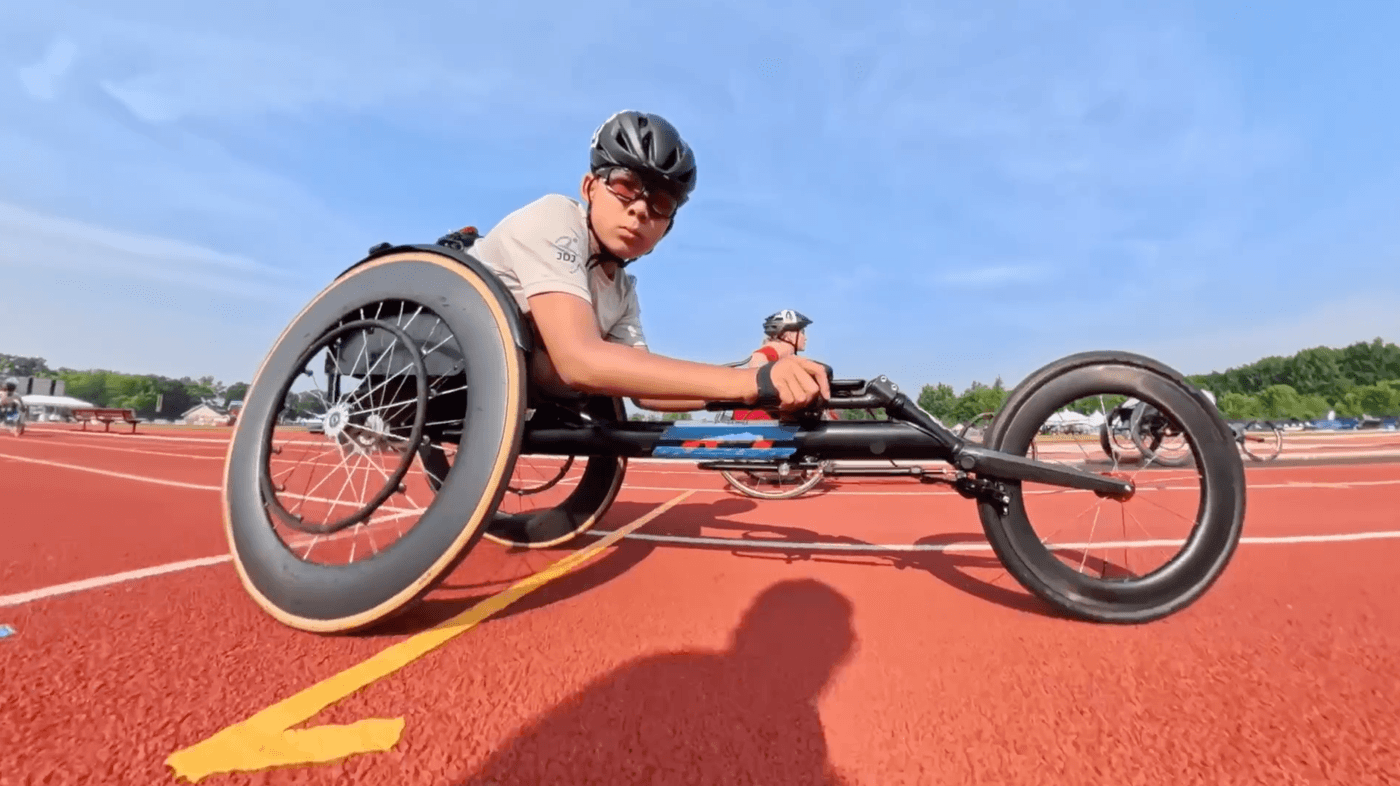 A person wearing a helmet and sunglasses competes in a track race using a recumbent handcycle on a red running track, with another racer visible in the background.