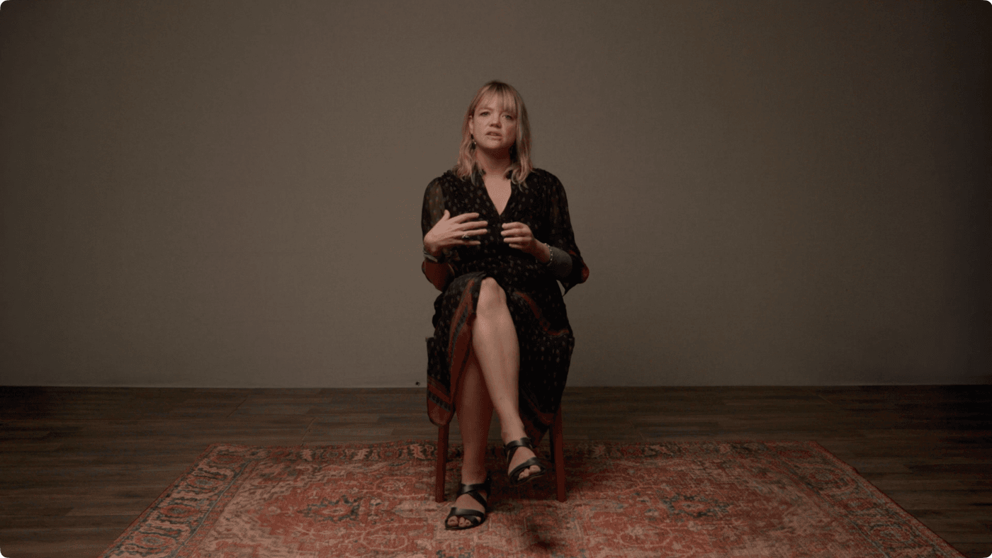 A blonde woman in a dark patterned midi dress sits on a wooden chair on an ornate rug, gesturing with her hands while speaking against a plain, light brown background.