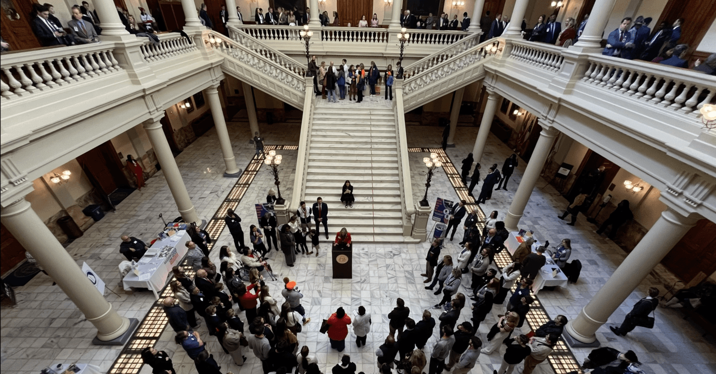 A high-angle view captures a crowd gathered in a large, marble-floored state capitol rotunda, where a woman stands at a wooden podium to address attendees while others observe from the grand staircase and surrounding balcony levels.