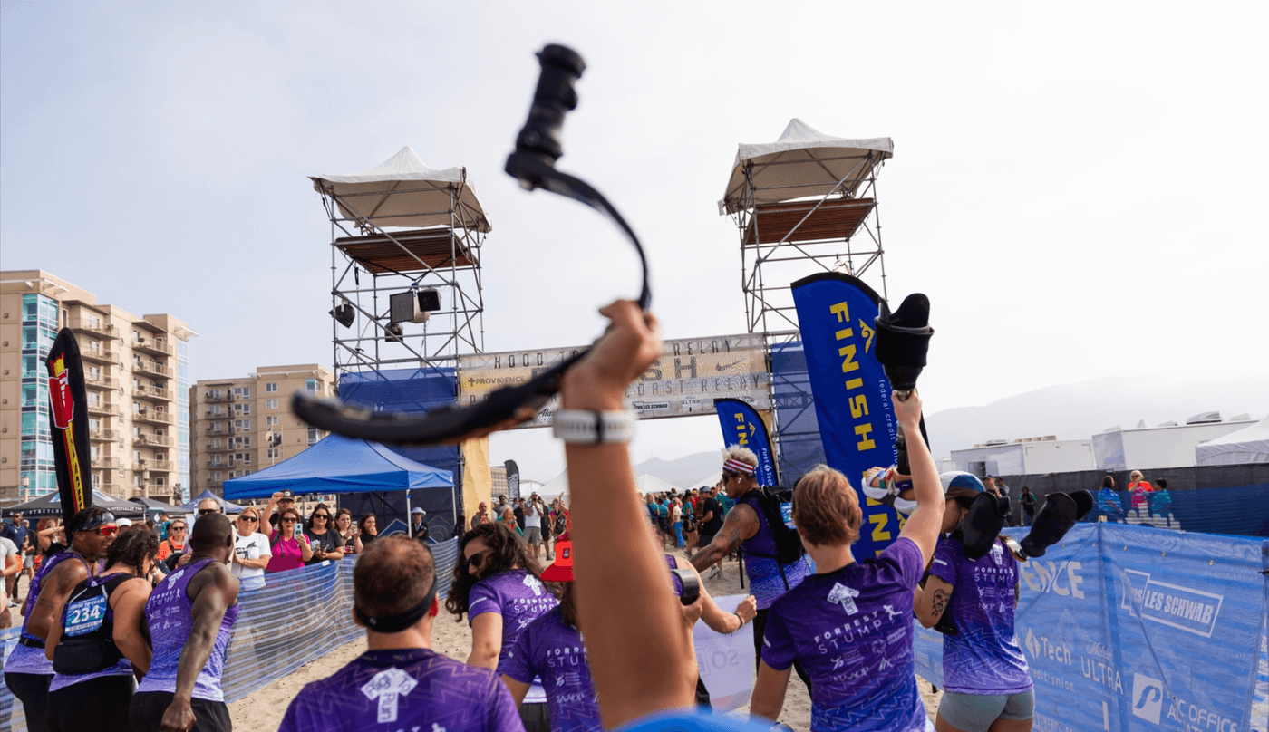 A person holds a prosthetic running blade high in the air while surrounded by teammates wearing purple "Forrest Stump" shirts at the finish line of the Hood to Coast relay race on a beach.
