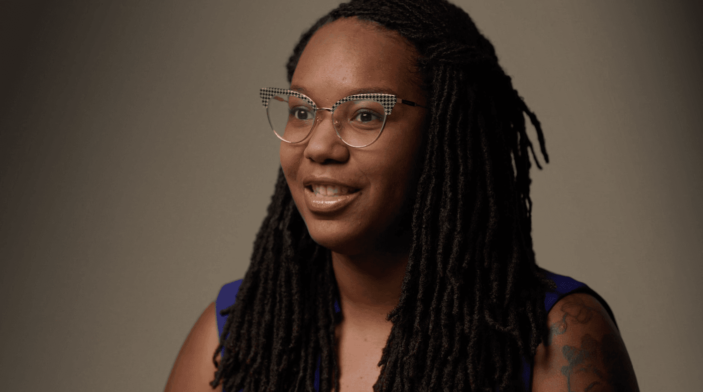 A woman with long, dark dreadlocks and glasses with a black-and-white patterned trim on the top corners looks slightly away from the camera while wearing a blue top against a neutral gray background.