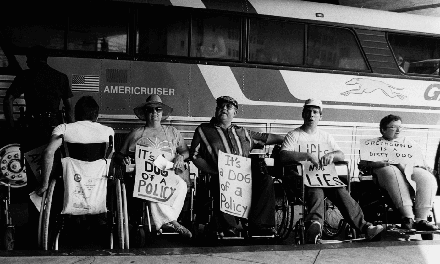 A black-and-white, low-angle photograph captures four protestors in wheelchairs lined up in front of an Americruiser bus, holding signs with slogans criticizing Greyhound, such as "It's a dog of a policy" and "Greyhound is a dirty dog."